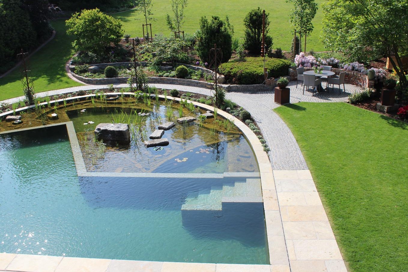 Ein Schwimmteich aus Naturstein und Kortenstahl mit eckigen und runden Formen und direkter Anbindung an die Terrasse. 