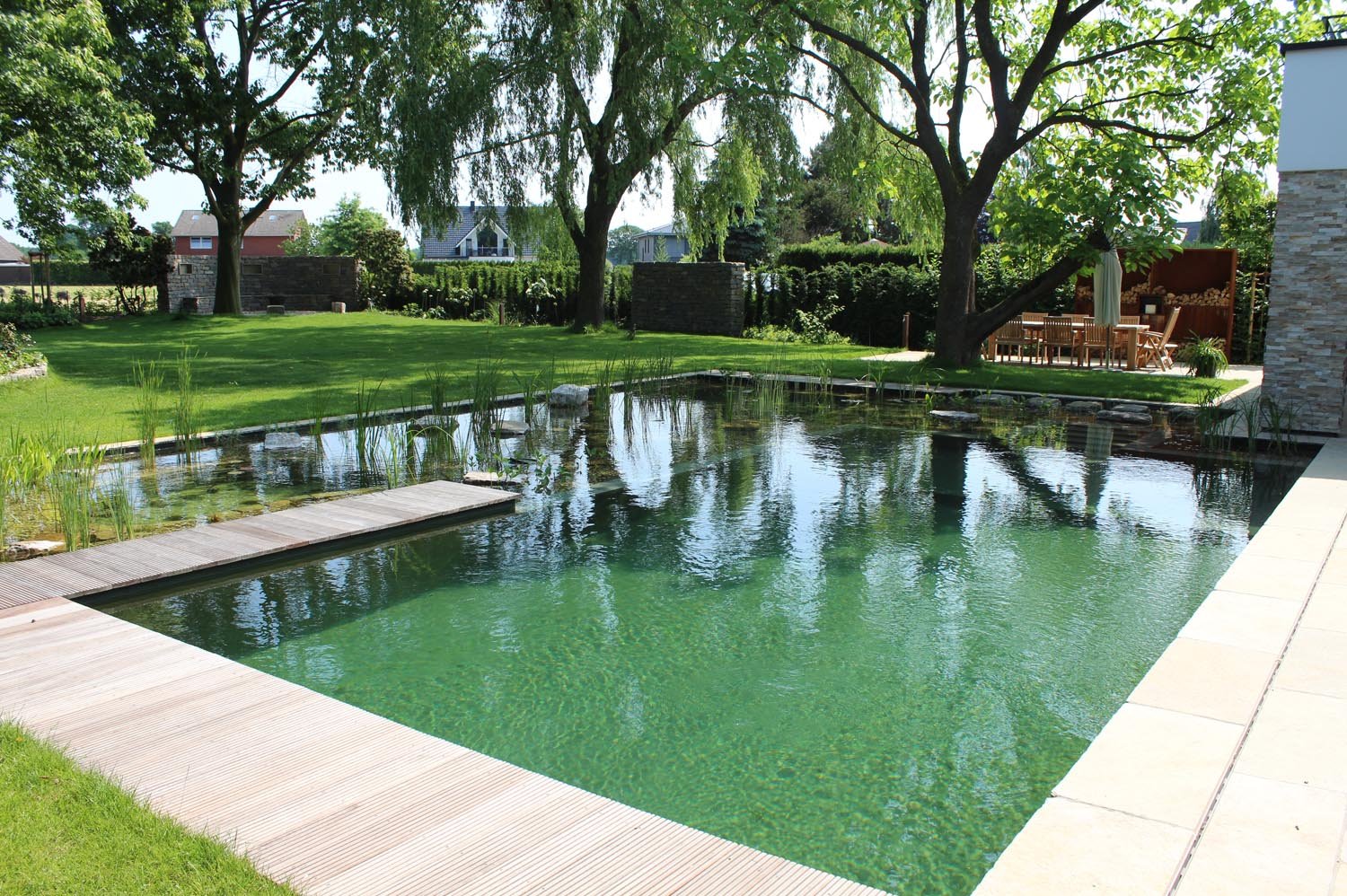 Design Schwimmteich aus Holz und Stein mit umliegender Terrasse und grünem Rasen. Wirkt harmonisch und verbunden. 