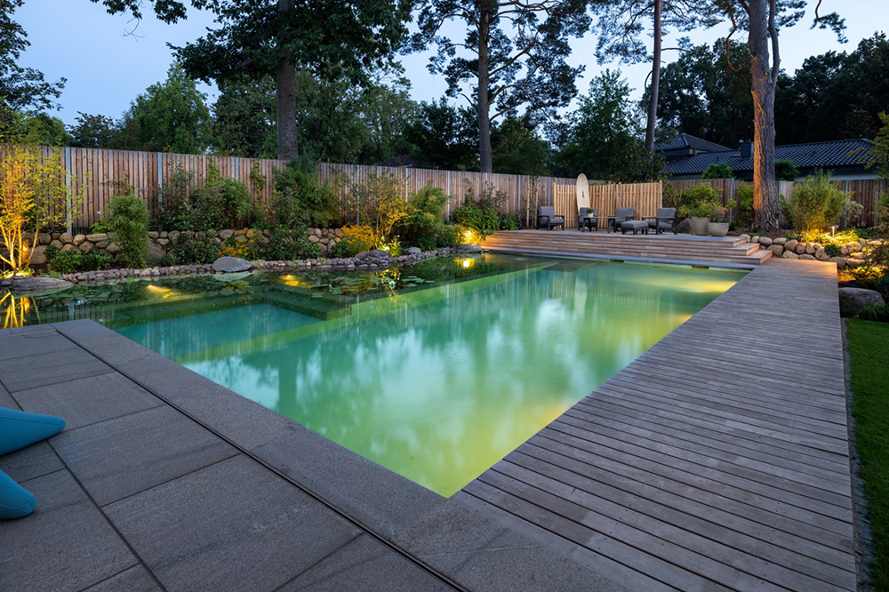 Ein Biotop Swimming Pond, gebaut von Garten Bitters, mit warmen Licht beleuchtet und Holzumrandung.
